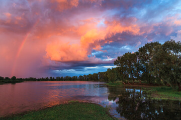 Rainbow and fiery orange clouds at sunset over the lake with reflection and green grass in the foreground