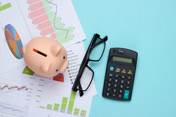 Piggy bank with graphs and charts, calculator, glasses on a blue background. Economic analysis,...
