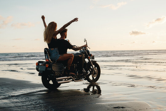 Young beautiful couple hipsters in stylish clothing for a retro motorcycle on the beach. Stylish couple roding motorbaki on black sand beach on balii, girl ith open arms feel freedom