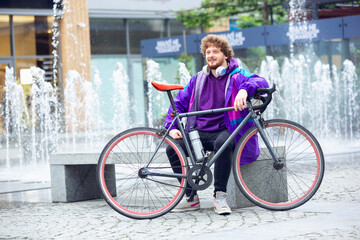 Fototapeta premium Handsome young man using headphones while sitting near his bicycle near fountain. Listen to music, scrolling news, chatting, talking with friends on the go. All the services in device anywhere.