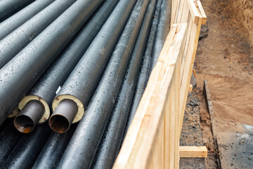 Heap of many new black insulated steel pipes at municipal construction site with wooden fencing around trench outdoors. Heating main district pipeline reconstruction. City development background