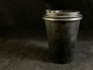 Paper cup of coffee with a lid on a black background. Disposable cup of coffee in the dark. Dishes for hot and cold drinks.