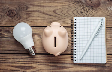 Saving electricity. Led light bulb and piggy bank, notebook on wooden background. Top view