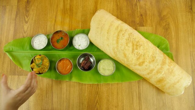 Woman hands serving a bowl of potato masala with South Indian cuisine Dosa. Top view of popular Dosa placed on a banana leaf with Sambhar  dosa filling  fried red chilies  and colorful chutneys