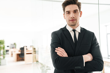 Image of serious young businessman wearing black suit standing with hands crossed