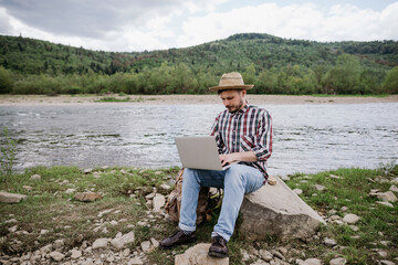 Young male hipster blogger is always online. A man sitting by the river and working on a laptop