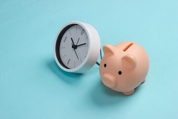 Time to invest. White clock and piggy bank on blue pastel background. Minimalistic studio shot.