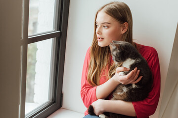 young woman holding cute cat while looking at window