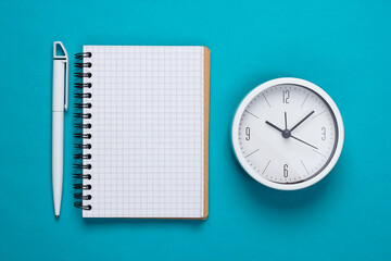 White clock and notebook on blue background. Minimalistic studio shot. Top view