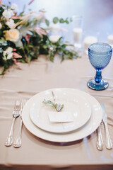 The table is covered with a white tablecloth, served. Plates, cutlery, napkin, blue glasses, seating cards, sprig of greens.