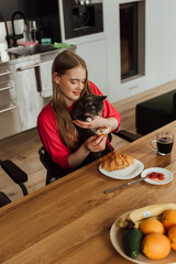 happy young woman holding tasty croissant near cute cat and cup of coffee