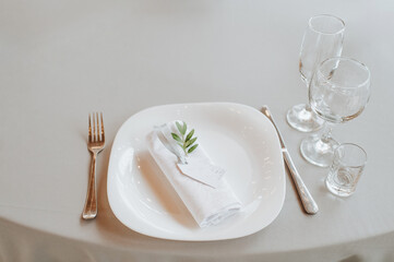The table is covered with a white tablecloth, served. Plates, cutlery, napkin, blue glasses, seating cards, sprig of greens.