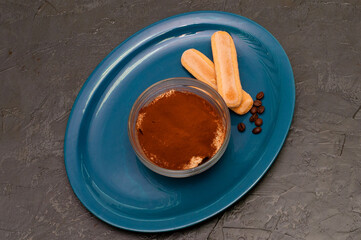 Dessert of italian cuisine tiramisu with cocoa and savoyardi cookies in a glass bowl on blue plate, gray concrete background, top view