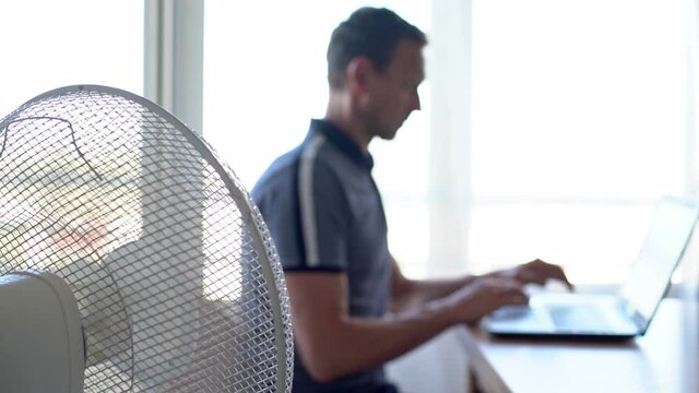 A man at a computer, laptop in a working environment, with the electric fan turned on. Blurred background.