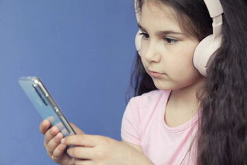 Cute child girl in headphones is using a smartphone, looking at camera and smiling on bright background. Listening music. Online talking.