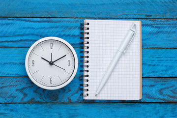 White clock and notebook on blue wooden background. Minimalistic studio shot. Top view