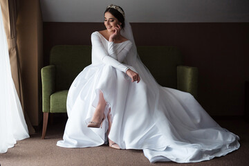 Beautiful bride in a luxurious dress, with makeup and hairstyle sitting on a sofa by the window. The bride is waiting for the arrival of the groom. Wedding morning