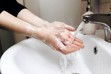 Two female hands with soapy foam in a stream of tap water in the bathroom close-up. The concept of hygiene, cleanliness and protection against coronavirus COVID-19.