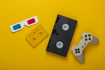 Anaglyph disposable paper 3d glasses, gamepad, video and audio cassette on yellow background. Retro media, entertainment 80s. Top view. Flat lay