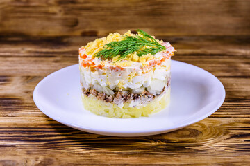 Plate with traditional russian salad Mimosa on a wooden table. Layered salad with potatoes, sardine, cheese, carrot, eggs and mayonnaise