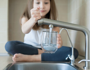 Little girl open a water tap with her hand holding a transparent glass. Kitchen faucet. Filling cup...