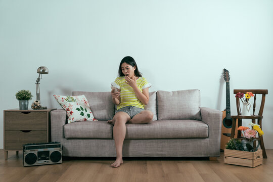 chinese woman shocked by bad news on cellphone. frowning young girl open mouth and cover by hand while looking at mobile phone screen on couch in home living room. surprised lady with text sms online