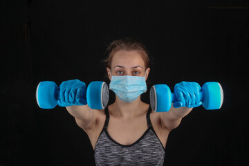 Fitness during quarantine from the covid-19 pandemic. Athletic woman In a medical mask and gloves holds a dumbbell in her hands on a black background.