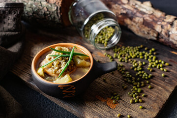 Soup with mash and potatoes. Decorated with green onions and peas of small beans from a jar. Serving in a ceramic pot on a charred wooden background with fabric and natural driftwood