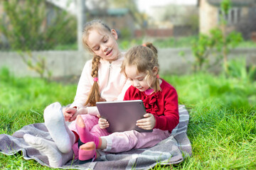 Smiling children girls sit on a blanket at the garden or park and playing with digital tablet together.