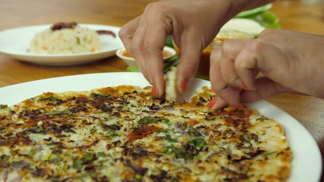 Closeup shot of woman hands eating South Indian vegetable Uttapam with sambhar. Group of South Indian foods like idli  uttapam  Upma  Vada served with potato masala  sambhar  and colorful chutneys