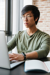 Image of handsome young asian man using laptop in apartment