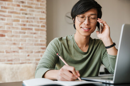 Image of asian man wearing eyeglasses talking on cellphone in apartment