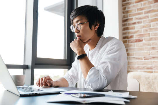 Image of young asian man using laptop while sitting at table in office