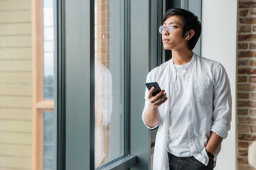 Image of young asian man wearing earphones using cellphone in office