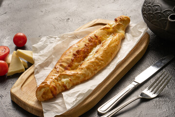 Khachapuri with cheese and tomatoes on a cutting wooden board next to cutlery, and ingredients on a black table.