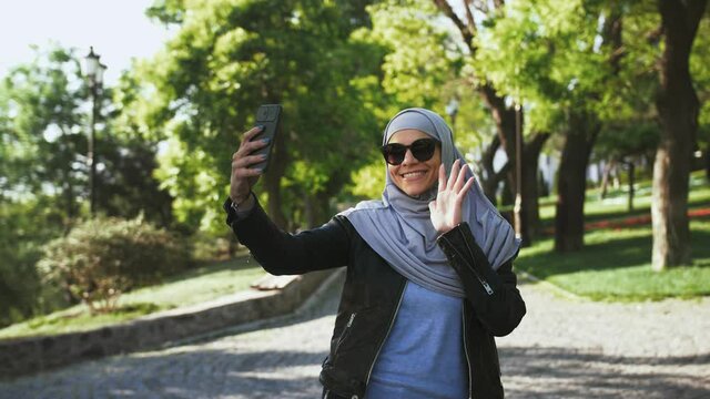 Muslim girl in sunglasses and hijab. She smiling, enjoying online video call on her mobile phone while walking in park with green trees. Slow motion