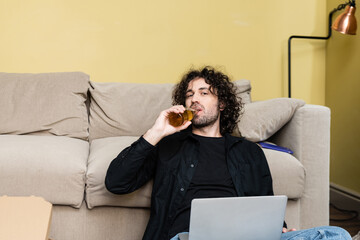 Fototapeta premium Handsome man looking at camera while drinking beer near laptop and pizza box at home