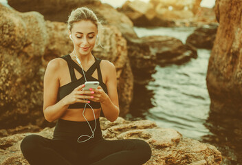 Young slim fit woman in sportwear listening to music in earphones and using smartphone while sitting on a stone at wild beach