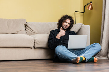 Low angle view of handsome man showing thumb up while working on laptop on floor