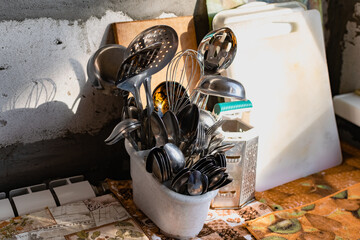 Metal Cutlery in a white stand. Silver spoons, forks, and stainless steel spatulas stand in the kitchen, with chopping boards and a grater at the back. Concrete gray walls with cement.