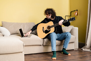 Curly man holding acoustic guitar and using laptop during online education in living room