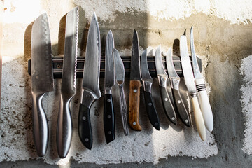 A set of metal knives of different sizes and shapes, hanging on a horizontal magnet holder. On a...
