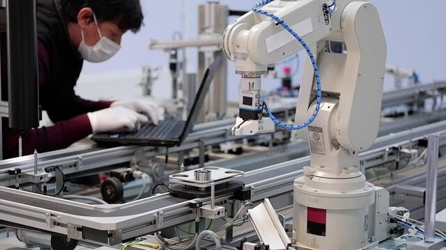 Close-up robot arm. Man with surgical mask and gloves against COVID-19 ( Coronavirusis ) working on laptop to programming smart factory automation and robot arm. industry 4.0 production line control.