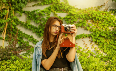 Young hipster woman in denim jacket and glasses enjoys film retro camera outdoors