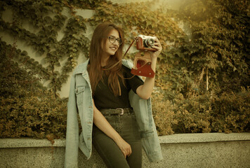 Young hipster woman in denim jacket and glasses enjoys film retro camera outdoors