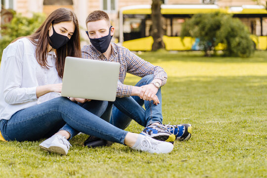 Ouple Students In Facial Masks Sitting On The Grass, Practicing Distance Learning, Using Laptop Computer, Beside Campus Building Outdoors