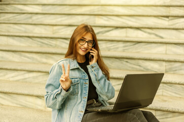 All in business. Young stylish woman in a denim jacket and glasses uses a laptop and talking on the phone while sitting on the stairs in the city. Remote work.