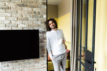 handsome man in pajamas holding coffee cup and looking at camera near blank lcd screen hanging on...