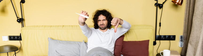 horizontal image of displeased man holding thumb down and looking at thermometer in bed