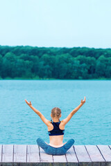 Back view of little girl is doing yoga exercise on grass of the shore of the lake.